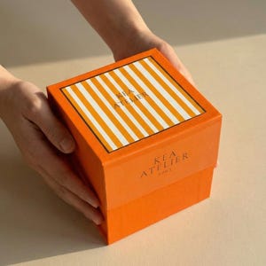 Hands gently holding and presenting a premium KEA ATELIER orange ribbed luxury gift box.