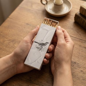 May include: A rectangular matchbox with a bird illustration is held in hands. The matchbox has a white background with a marble-like texture. The bird is perched on a branch. The match heads are visible inside the open box.
