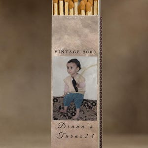 May include: A rectangular matchbox with a vintage design. The top section holds matches with brown tips. The box features a photo of a child, the text "VINTAGE 2003", and "Diana's Turns 23". The box has a brown and beige color scheme.