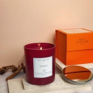 A stylish KEA ATELIER minimalist red glass jar soy candle displayed elegantly next to its signature orange ribbed luxury gift box.
