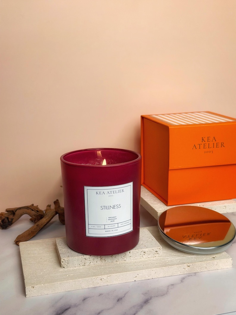A stylish KEA ATELIER minimalist red glass jar soy candle displayed elegantly next to its signature orange ribbed luxury gift box.
