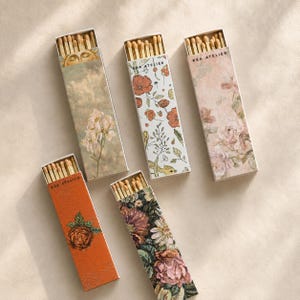 May include: Five rectangular matchboxes with wooden matches, each featuring unique floral or celestial designs. The boxes display the text KEA ATELIER and showcase various patterns including a sun, orange poppies, pink roses, a single rose, and a dark floral bouquet.