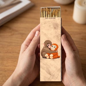 May include: A matchbox held by hands, featuring a cartoon illustration of two hugging monkeys. The box is tan with the text "KEA ATELIER" at the top. The matches are visible inside the open box.