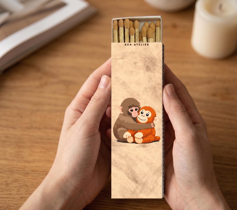 May include: A matchbox held by hands, featuring a cartoon illustration of two hugging monkeys. The box is tan with the text "KEA ATELIER" at the top. The matches are visible inside the open box.
