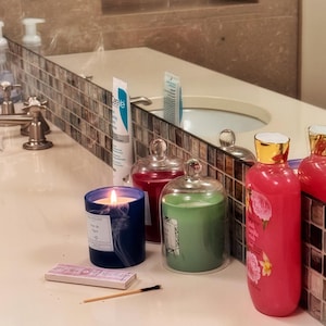 May include: A lit blue candle, a green candle in a glass cloche, and a pink bottle with floral designs sit on a bathroom counter. A box of matches and a burnt matchstick are also present. A tube of CeraVe is visible.