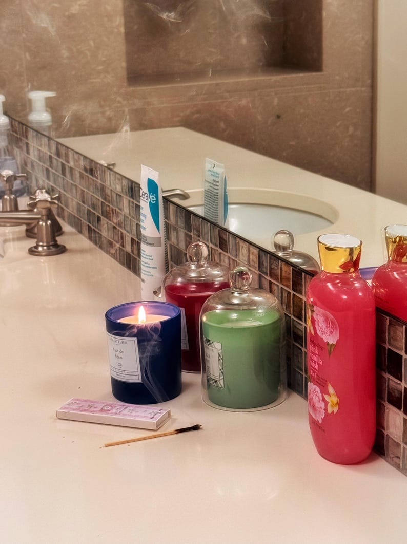 May include: A lit blue candle, a green candle in a glass cloche, and a pink bottle with floral designs sit on a bathroom counter. A box of matches and a burnt matchstick are also present. A tube of CeraVe is visible.
