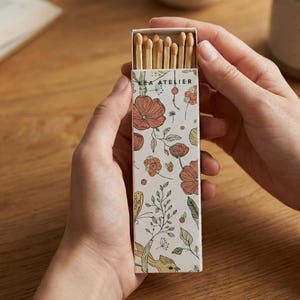 May include: A rectangular matchbox held by hands, featuring a floral design with orange and green flowers and the text "KEA ATELIER". The box contains several wooden matches with brown tips.