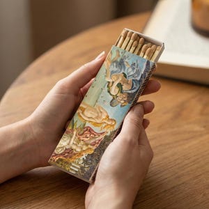 May include: A rectangular matchbox featuring a Renaissance painting design, held in hands. The matchbox is open, revealing the wooden matchsticks inside. The artwork depicts figures in shades of blue, beige, and red.