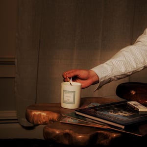 May include: A lit candle in a white ceramic container with a label, placed on a wooden table. The candle is being lit by a match. Several books and a matchbox are also on the table. The scene is dimly lit, creating a warm and inviting atmosphere.