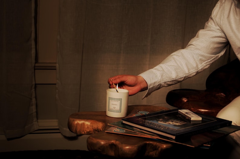May include: A lit candle in a white ceramic container with a label, placed on a wooden table. The candle is being lit by a match. Several books and a matchbox are also on the table. The scene is dimly lit, creating a warm and inviting atmosphere.
