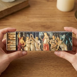 May include: A rectangular matchbox featuring a Renaissance-style painting of figures in a forest setting. The matchbox is held open, revealing the wooden matchsticks inside. The artwork depicts several figures in various poses, with a color palette of muted tones.