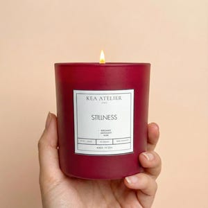 A hand elegantly holding a KEA ATELIER bold red glass jar soy candle, representing a luxurious masculine home decor gift.