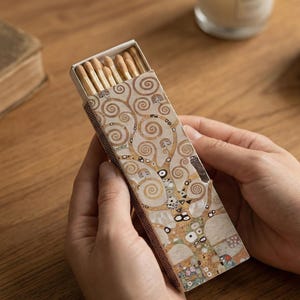 May include: A decorative matchbox featuring a detailed, swirling tree design in gold and brown tones. The matchbox is held open, revealing the wooden matchsticks inside. The design is reminiscent of the artwork of Gustav Klimt.