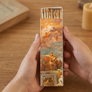 May include: A rectangular matchbox with a classical painting design. The box features the text "KEA ATELIER" at the top. The artwork depicts figures in a cloudy sky and a warrior with a shield. The match heads are visible at the top.