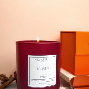 A close-up of the masculine Bergamot Mahogany Musk red jar candle resting on a surface with the orange gift box in the warm background.