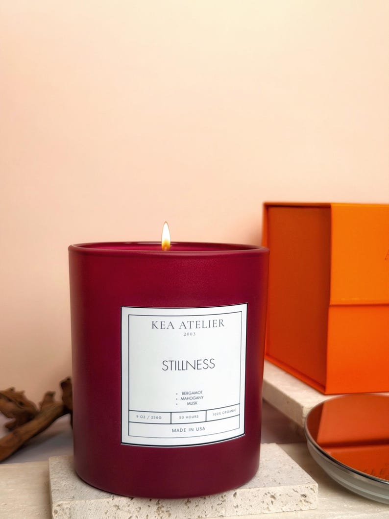 A close-up of the masculine Bergamot Mahogany Musk red jar candle resting on a surface with the orange gift box in the warm background.