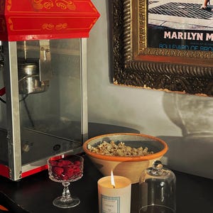 May include: A red and silver popcorn machine stands next to a bowl of popcorn, a lit candle, and a glass of raspberries. A framed Marilyn Monroe print hangs on the wall. A glass cloche and a matchbox are also on the table.