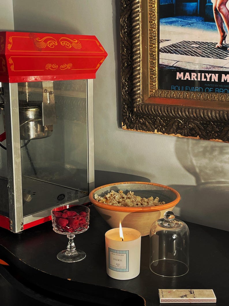 May include: A red and silver popcorn machine stands next to a bowl of popcorn, a lit candle, and a glass of raspberries. A framed Marilyn Monroe print hangs on the wall. A glass cloche and a matchbox are also on the table.