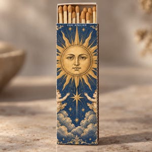 May include: A rectangular matchbox featuring a dark blue background with a gold sun face illustration, two cherubs reaching toward a central star, and clouds. The text KEA ATELIER is printed at the top. The box is filled with wooden matches.