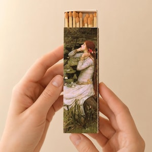May include: A rectangular matchbox featuring a reproduction of a painting of a person with long red hair in a white dress sitting by a pond. The box is partially open at the top, revealing a row of wooden matches with light brown tips.