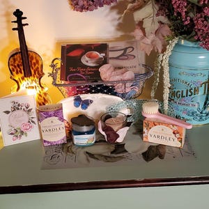 May include: A still life arrangement featuring a teal blue tea tin with the text "Traditional English Tea", a small glass jar with a silver lid, a white and blue butterfly design eye mask, a small white card with the text "Thank You", a small purple box with the text "Yardley English Lavender", a small pink box with the text "Yardley Milk & Buttercups", a small glass jar with a silver lid, a small white plate with a blue butterfly design, a small basket with a book titled "Tea-Time Serenade Tea Tunes", a small pair of silver scissors, and a small wooden brush.