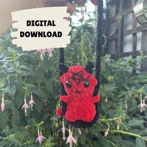 May include: A handmade red plush devil doll with black horns and a pentagram on its head, suspended by a black cord. The doll is set against a backdrop of green foliage and pink flowers. The words "DIGITAL DOWNLOAD" are in a white box.
