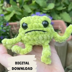 May include: A light green crochet frog with black eyes and a stitched mouth. The frog is held in a hand, with the text "DIGITAL DOWNLOAD" overlaid on the image. The frog has a soft, plush texture. The background shows foliage.