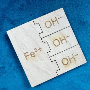 May include: A wooden model of the chemical formula for iron hydroxide, Fe(OH)3. The model is made of three separate pieces, each with the chemical formula written on it. The pieces are connected by a series of interlocking tabs.