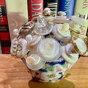 Op de afbeelding: Een witte theepot van keramiek met een bloemenpatroon is gevuld met een boeket van witte en gouden knopen.
