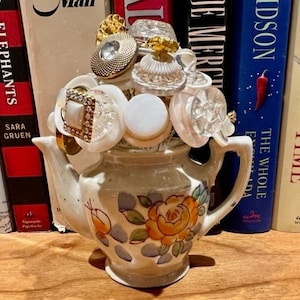 Puede incluir: Una tetera blanca con un diseño floral está llena de botones y otros artículos decorativos. La tetera está sentada sobre una superficie de madera frente a una estantería.