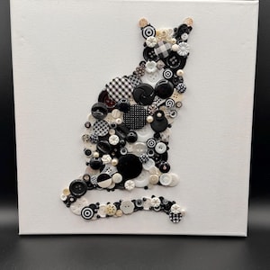 May include: A wall art piece featuring a cat silhouette created from a mosaic of various black, white, and cream-colored buttons arranged on a square white canvas. The buttons vary in size, texture, and pattern, including polka dots and checks.