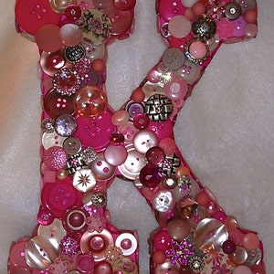 May include: A decorative letter "K" crafted from various shades of pink buttons. The buttons are of different sizes, shapes, and textures, including pearlized, floral, and metallic accents. The letter is a vibrant, handmade piece.