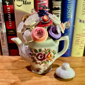 Puede incluir: Una tetera blanca con un diseño floral y una tapa, llena de un ramo de botones coloridos. La tetera está sentada sobre una superficie de madera frente a una estantería.