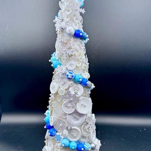 May include: A conical decorative tree sculpture covered in white buttons, blue and white beads, and silver glittery snowflake ornaments. The structure is topped with a clear, pointed star ornament, creating a textured, layered appearance.