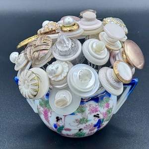 May include: A collection of assorted buttons in shades of white, cream, gold, and metallic tones arranged in a bouquet style. The buttons are mounted on thin wires and displayed inside a small, round, white ceramic bowl with blue handles.