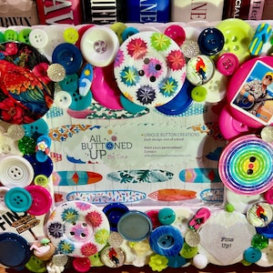 May include: A white picture frame decorated with colorful buttons, featuring a surfboard and palm tree design. The frame includes a business card for "All Buttoned Up by Tina" with the text "Unique Button Creations, Each design, one of a kind".