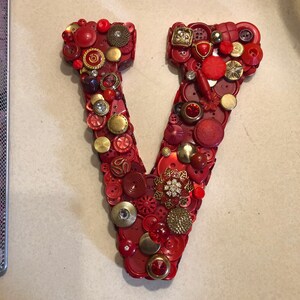 Könnte beinhalten: Ein roter Buchstabe V aus Knöpfen. Die Knöpfe sind größtenteils rot, mit einigen goldenen und ein paar klaren oder glitzernden Knöpfen.