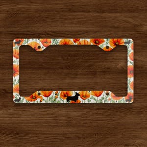 May include: License plate frame with a floral poppy design in orange and green on a white background. Black silhouettes of a dog are interspersed throughout the design. The frame is rectangular with cutouts for mounting.