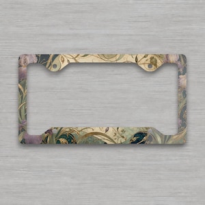 May include: A rectangular license plate frame featuring a decorative botanical pattern with muted purple flowers, dark teal circular accents, and flowing gold-toned vines against a cream and dark background with four mounting holes.