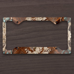 May include: A license plate frame with a brown leather-like background and white floral and leaf designs. The frame has turquoise and pearl-like accents. The frame is rectangular with rounded corners and four mounting holes.