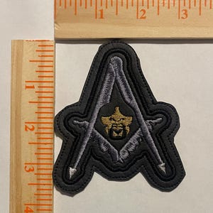 May include: Black embroidered patch featuring a gold emblem within a square and compass design. The patch is approximately 2.5 inches in height and width. The patch has a black border and is on a white surface.