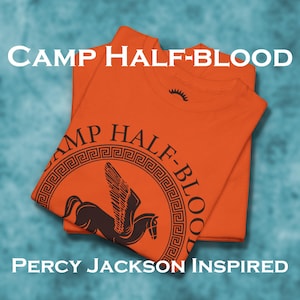 Könnte beinhalten: Zwei übereinander gestapelte orangefarbene T-Shirts. Das obere T-Shirt zeigt eine schwarze Grafik eines geflügelten Pferdes in einem Kreis mit dem Text "Camp Half-Blood" darum herum. Das untere T-Shirt hat den Text "Percy Jackson Inspired" aufgedruckt.