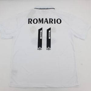 Puede incluir: Camiseta de fútbol blanca con el nombre "ROMARIO" impreso en negro en la parte superior de la espalda. El número "11" se muestra en negro en la espalda, con dos rayas verticales negras y detalles de estrellas. La camiseta tiene cuello y mangas cortas.