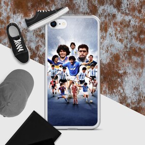 Op de afbeelding: Een doorzichtige telefoonhoes met een collage van voetballers in verschillende tenues. Een zwarte pet, zwarte sneakers en een zwarte portemonnee liggen ernaast. De achtergrond is getextureerd en roestig.