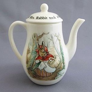 May include: A white ceramic teapot with a lid featuring a Beatrix Potter illustration of Mrs. Rabbit in a red hooded coat carrying a basket. The teapot has a handle and a spout.