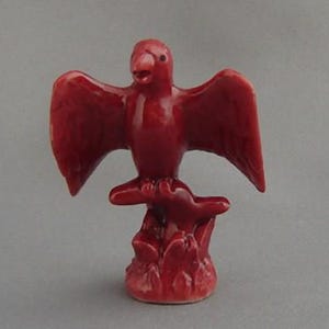 May include: A red ceramic figurine of a bird perched on a branch. The bird has its wings spread out and its head tilted to the side. The figurine is painted in a solid red colour.