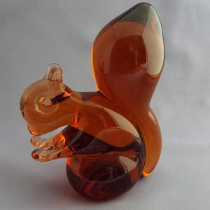 May include: A decorative glass squirrel figurine in shades of amber and brown. The squirrel is posed in a sitting position, with its tail arched upwards. The figurine is made of transparent glass, allowing light to pass through.