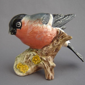 May include: A ceramic bullfinch ornament perched on a branch. The bird features a black head, a red breast, and grey wings. The branch is brown with yellow floral details. A decorative item, likely for display.