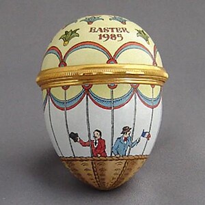 May include: A white porcelain egg-shaped trinket box with a gold lid. The box is decorated with a hot air balloon scene and the text "Easter 1985".