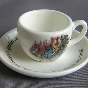 May include: A white teacup and saucer set with a Peter Rabbit illustration. The saucer has the text "Peter Rabbit's garden, a treasure" printed on it.